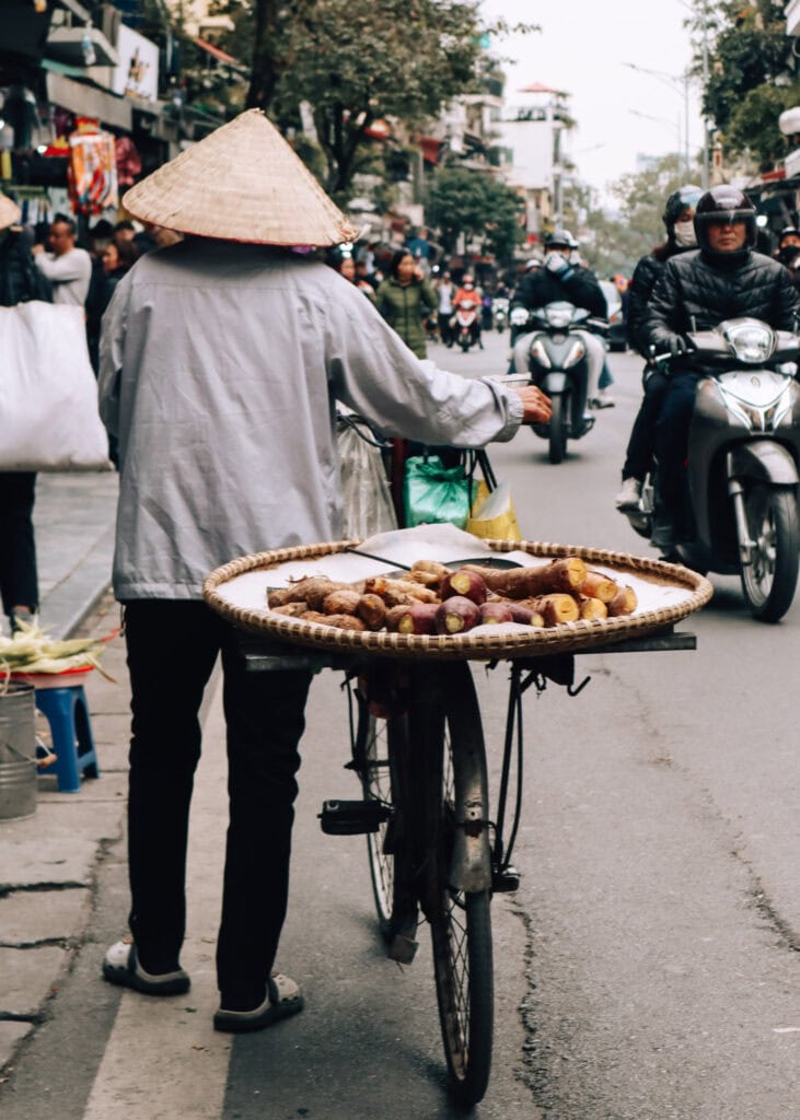 A street vendor in Hanoi walking a bike with root vegetables on a basket.