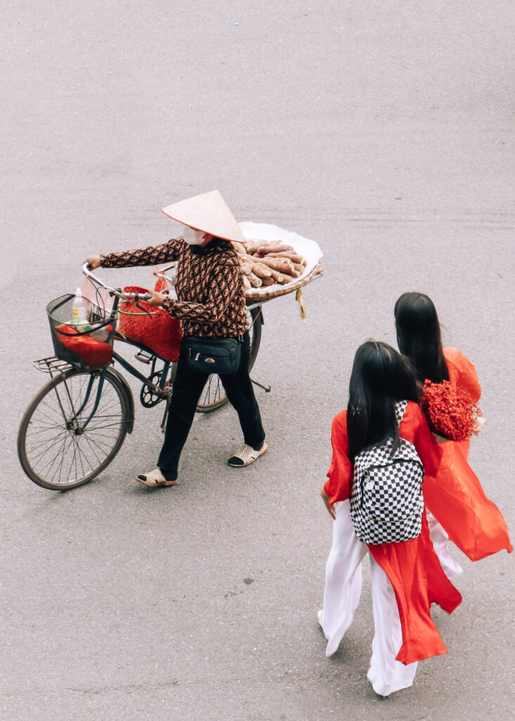 Two girls with long black hair and red, traditional dresses walking next to a street seller with a bike in Hanoi.