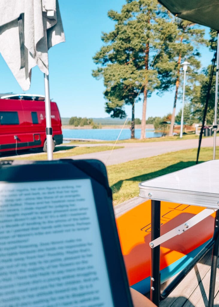Someone holding a Kindle in front of a camping table, a SUP and campers by a lake.