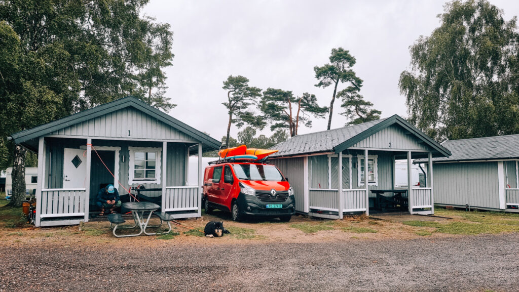 Man and a dog in front of a camping cabin in Sweden. Red van with kayaks on the roof parked between two cabins.