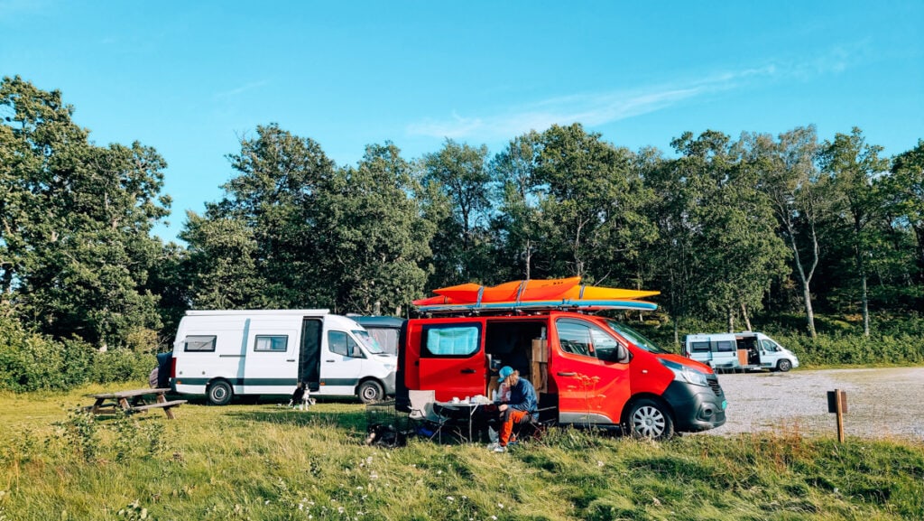 A man sitting by a camping table in front of a red van. Several campers behind.