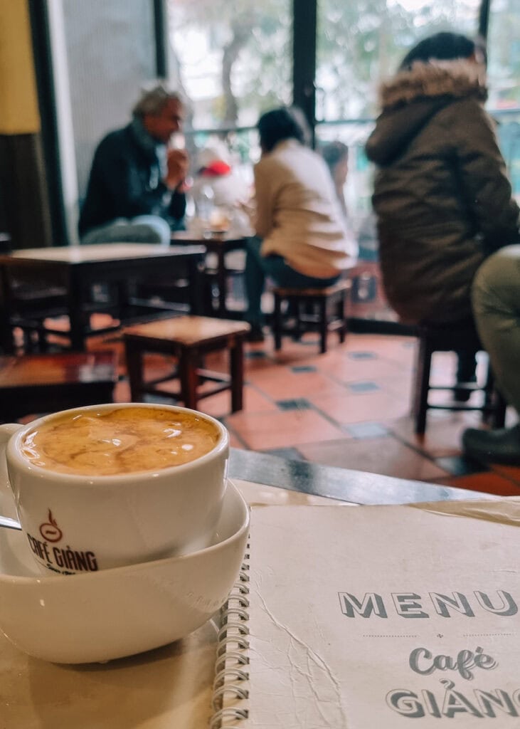 A cup of egg coffee sitting on a table at Café Giang. People seen sitting on stools in the background.