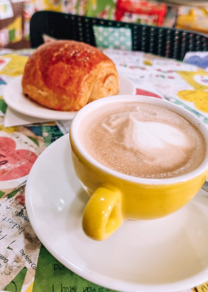 A yellow cup with coconut coffee next to a croissant at The Note Coffee.