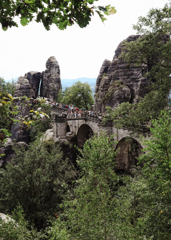 The Bastei Brücke seen through lush, green forest. Lots of people standing on the bridge.