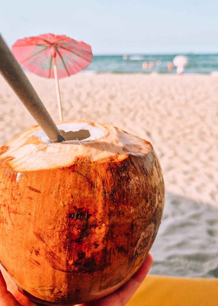 A coconut with straw and umbrella in front of a beach with the sea in the background.