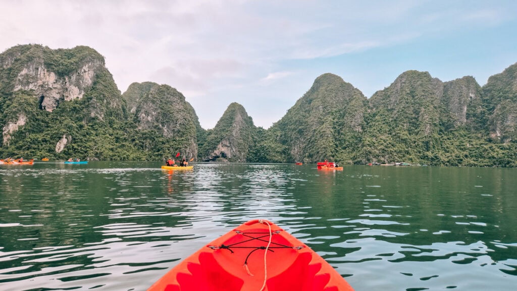 A picture taken from a kayak, seen at the bottom, with several kayaks in front of typical mountains in Halong Bay.