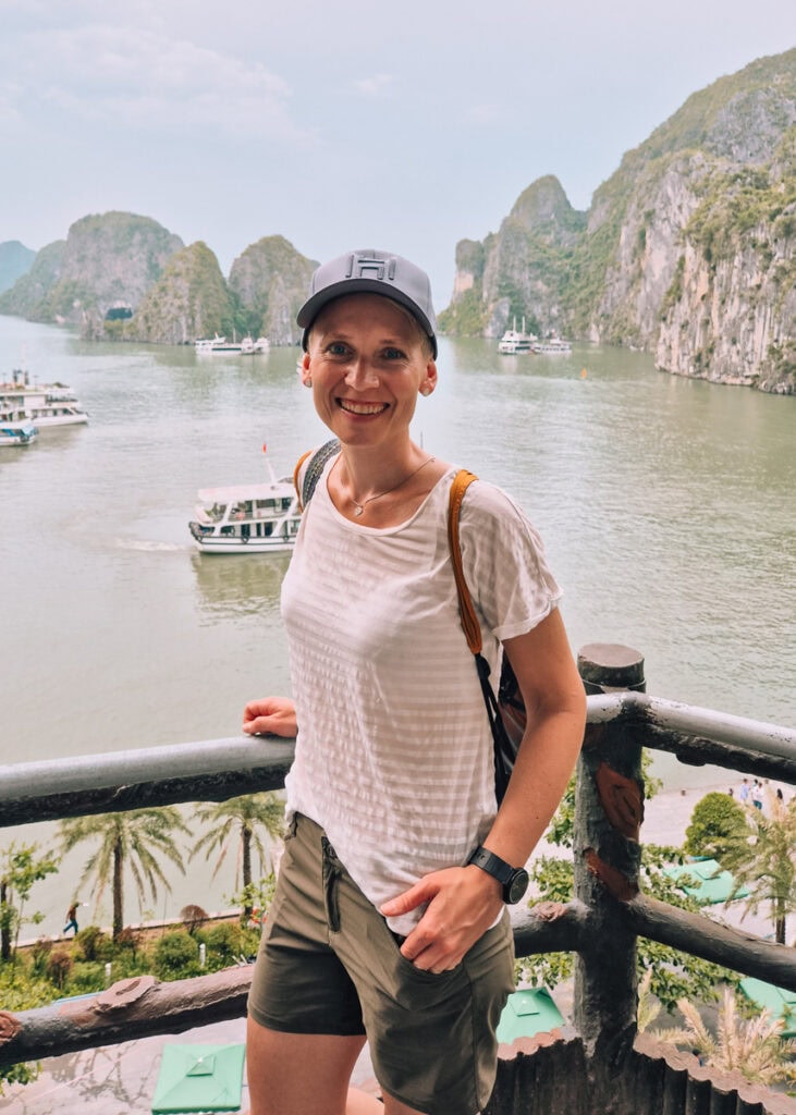 Anne (me) wearing cap, t-shirt and shorts smiling at the photographer by the entrance to the Sung Sot Cave with Halong Bay in the background.
