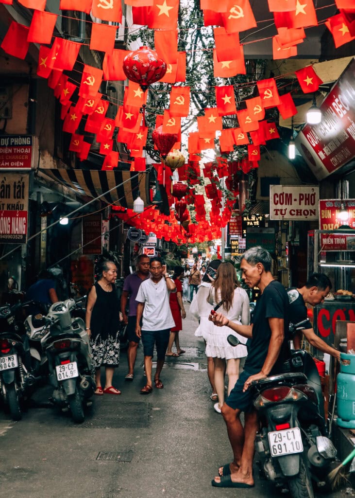 Street scene in Hanoi. Lots of small red flags and lanterns hanging from above, people going on with daily life.