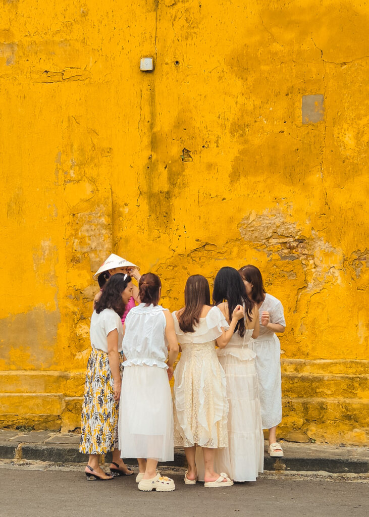 Six girls dressed in white seen from behind while they look at a phone next to a yellow wall.