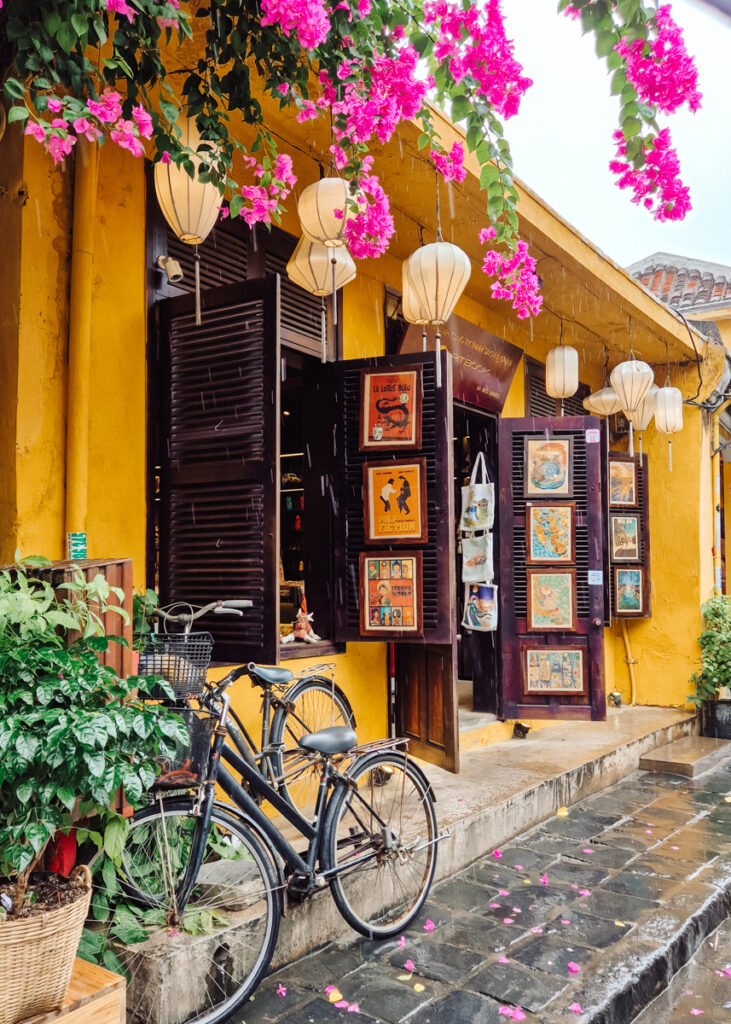 Two bicycles parked outside a shop. Yellow walls and posters hanging on the window frames and door, lanterns above.