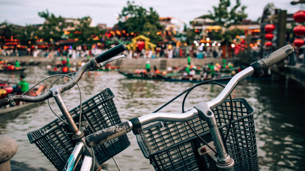 Two bike handlebars seen infront of the river in Hoi An, lots of boats with lanterns can be spotted behind.