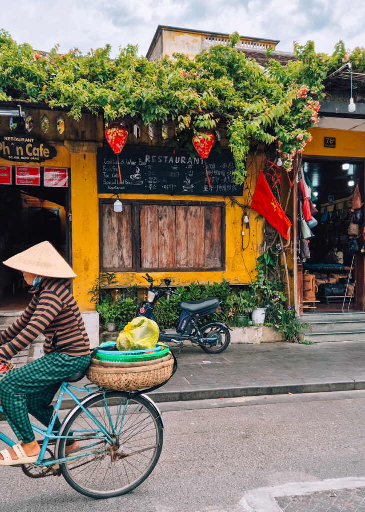 Street scene in Hoi An. A woman in a conical hat cycling past a yellow-painted café.