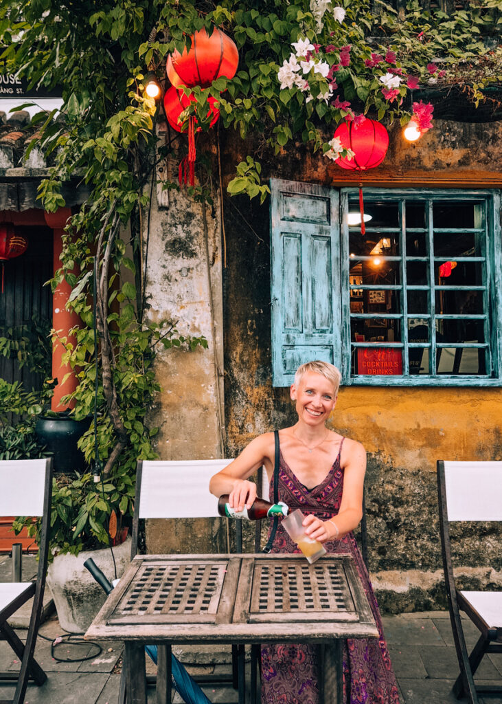 Anne (me) smiling while pouring beer on a table outside a restaurant in Hoi An. Flowers and lanterns hanging from the roof, yellow wall and blue windos seen behind.