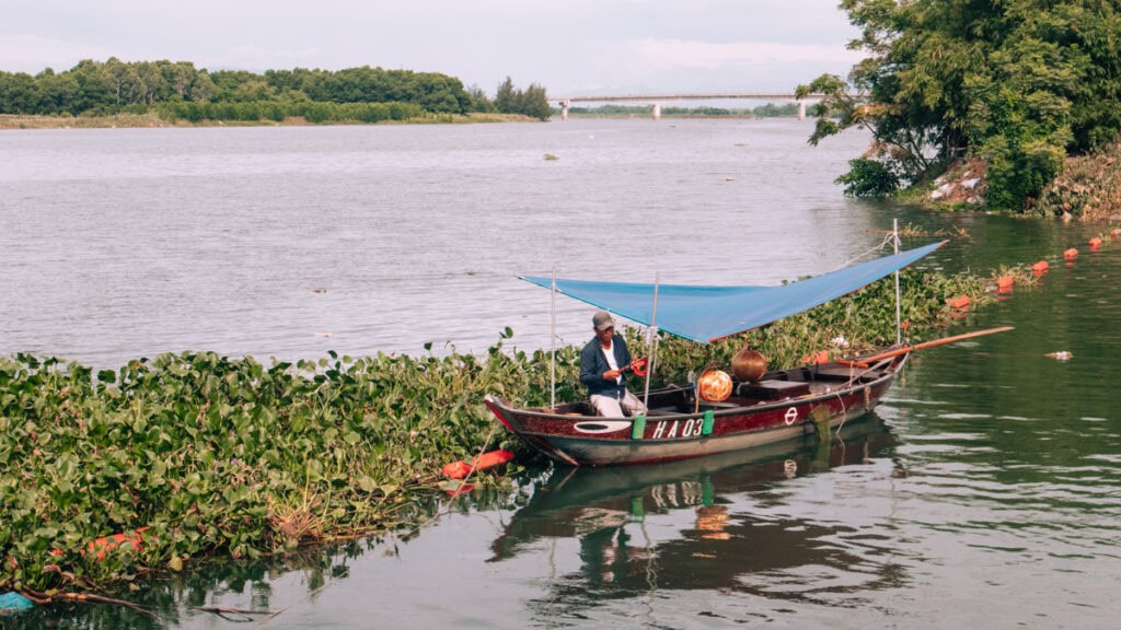 A fisherman sitting under a tarp in his wooden boat next to water lillies on the river outside Hoi An.
