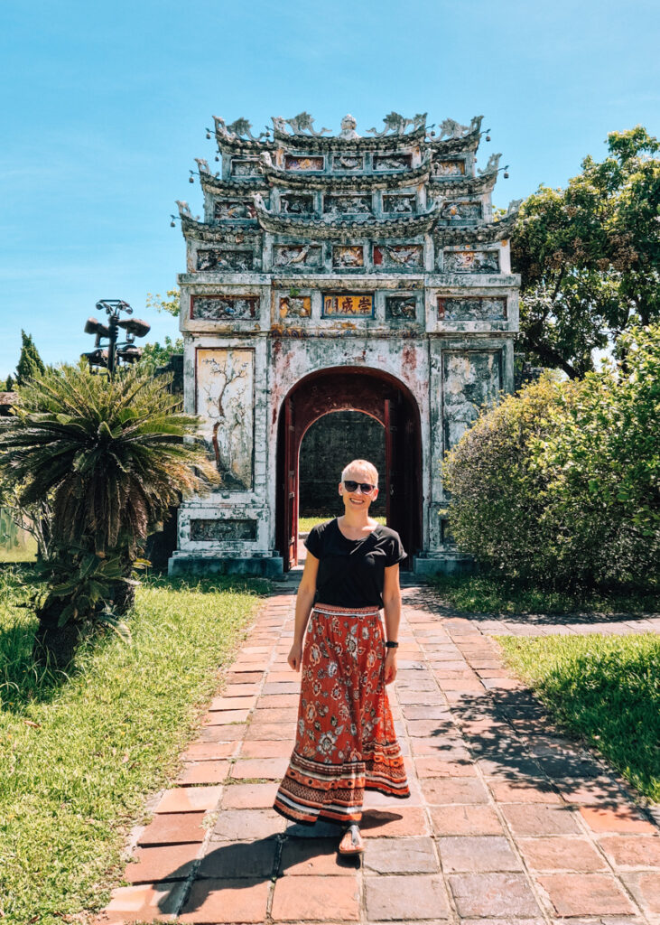 A woman wearing a patterned, orange skirt walking in front of an old gate inside the Imperial Citadel in Hue.