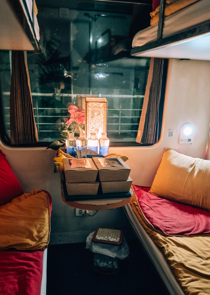 Interior of a Lotus Train compartment in Vietnam. Two bunk beds with red and yellow linen and a table with lamp and breakfast boxes and water for 4 people.