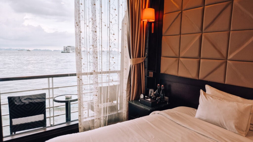 Interior of a cabin on a MilaLux Cruise. Half of a double bed next to a chest of drawers and balcony outside large windows.