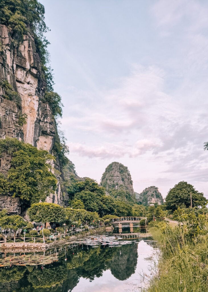Landscape in Ninh Binh with limestone karsts mirroring in a river. A small bridge and a garden behind a wooden fence.