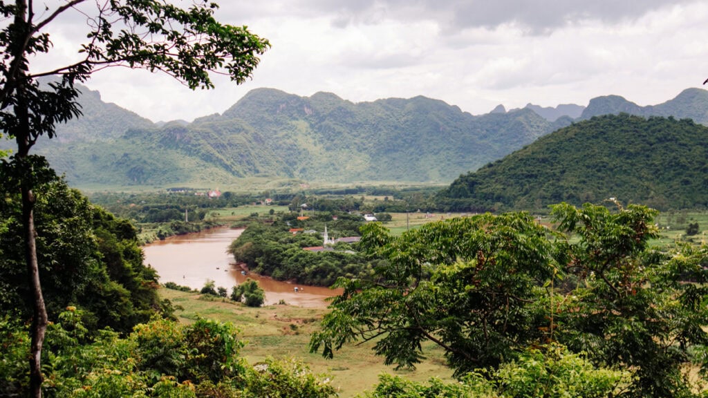Landscape view from the trail to Tien Son Cave. The brown, winding river, a small settlement with a church and foresty mountains.