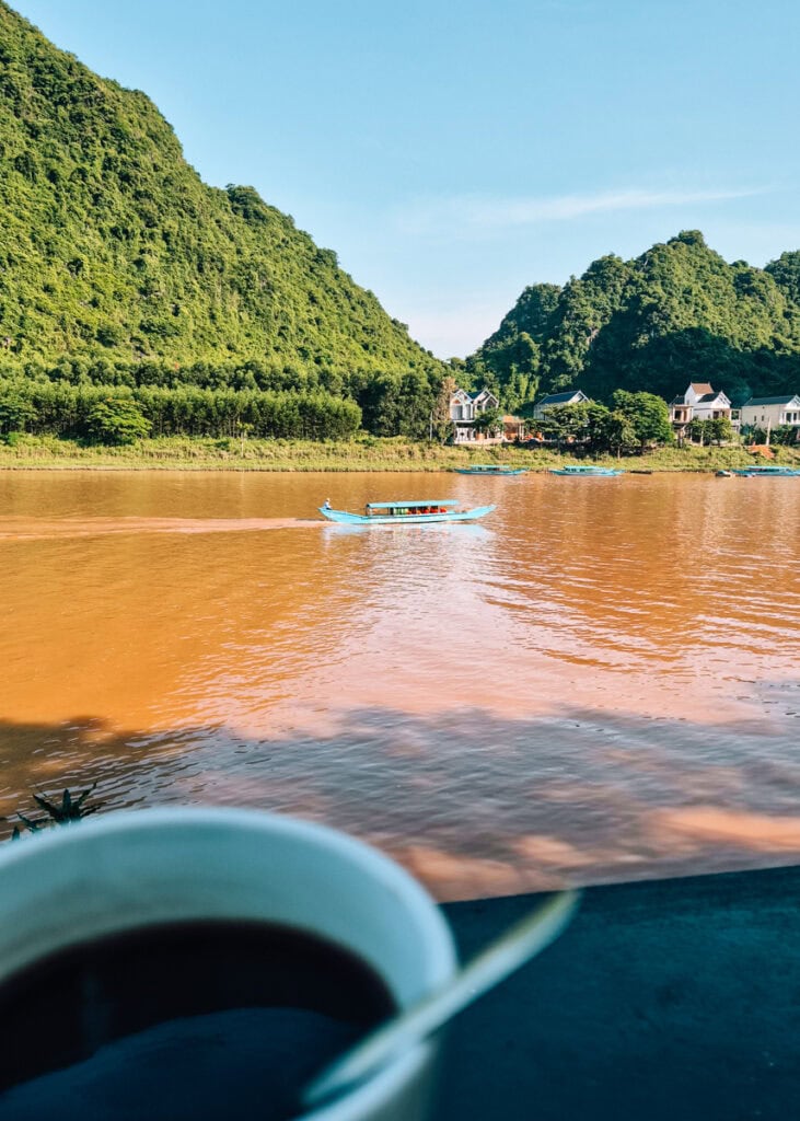A cup of coffee in front of the Phong Nha river. A blue boat sailing with forest-covered mountains behind.