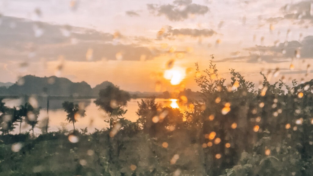Sunrise over a lake outside Dong Hoi in Vietnam. Greenery in front and small, foresty mountains behind. All seen through a train window with rain drops.