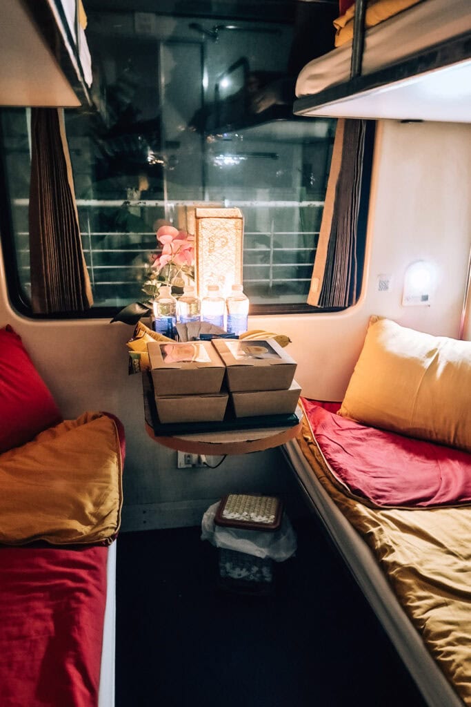 Interior of a 4-bed compartment on the Lotus Train. Yellow and red bedding, a table with 4 food boxes, water and snacks in addition to a lamp and a pink flower.