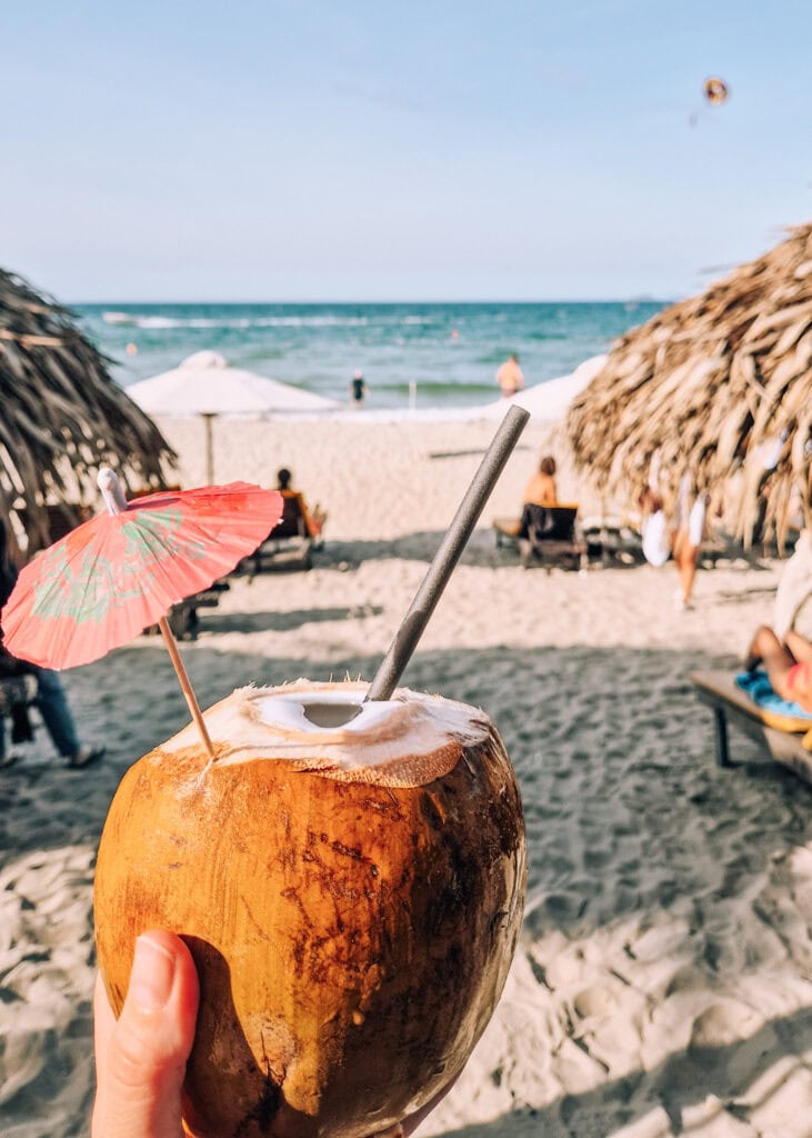 A hand holding a coconut in front of umbrellas and the sea.
