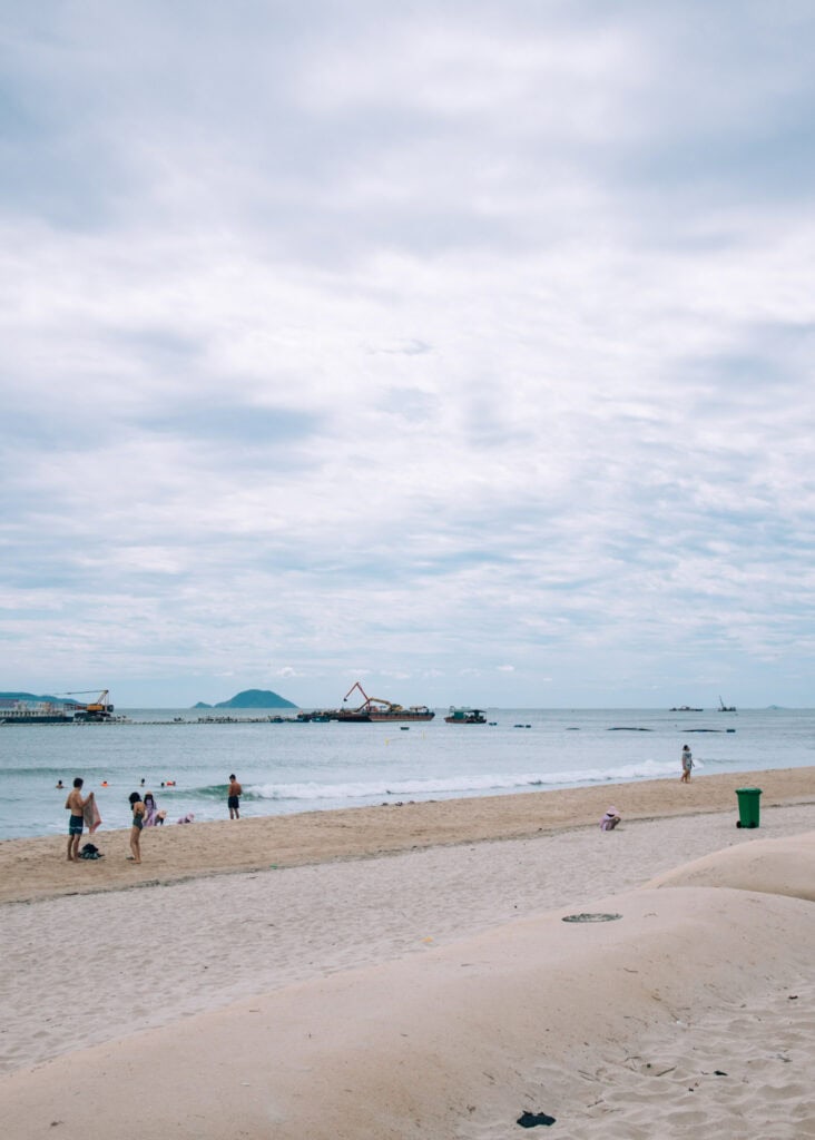 People swimming at Cua Dai Beach. Large boats and machines working on the beach in the distance.