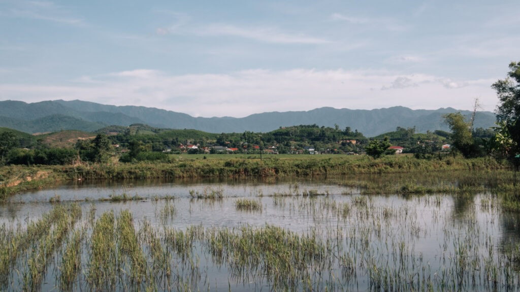 Landscape view of the valley near Phong Nha Farmstay. A sall pond in front of green fields, clusters og houses and mountains in the back.