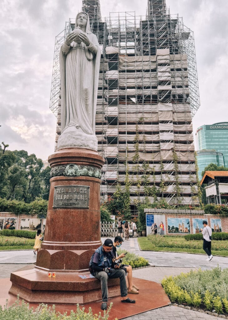 The Notre-Dame Cathedral in Saigon behind scaffolding. A statue in front, with two men sitting and looking at their phones.